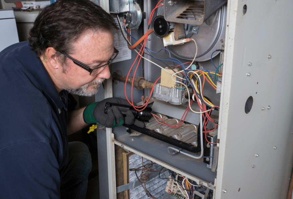 A technician wearing safety glasses and gloves inspects and repairs the interior components of a furnace or heating system, using a flashlight to examine the burner and wiring assembly.