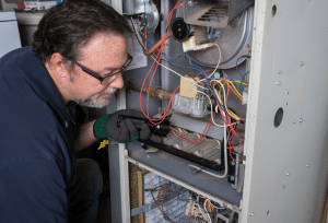 A technician wearing safety glasses and gloves inspects and repairs the interior components of a furnace or heating system, using a flashlight to examine the burner and wiring assembly.